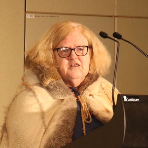 Image of Professor Di Kerr. She is standing behind a podium and wearing glasses and a dark blue shirt underneath a possum skin cloak.
